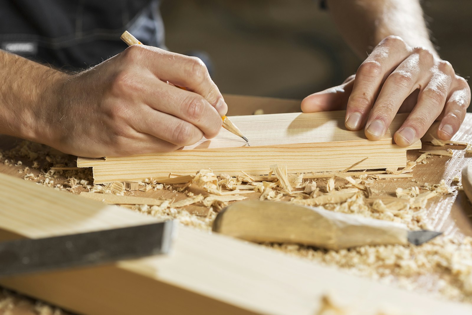 Hände eines Handwerkers, der an der Werkbank eine Linie auf ein Holzbrett zeichnet.